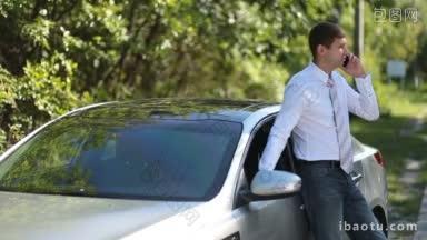 英俊的年轻商人打着领带，靠在停着的汽车上，用智能手机聊天，背景是农村风景，成功的企业家